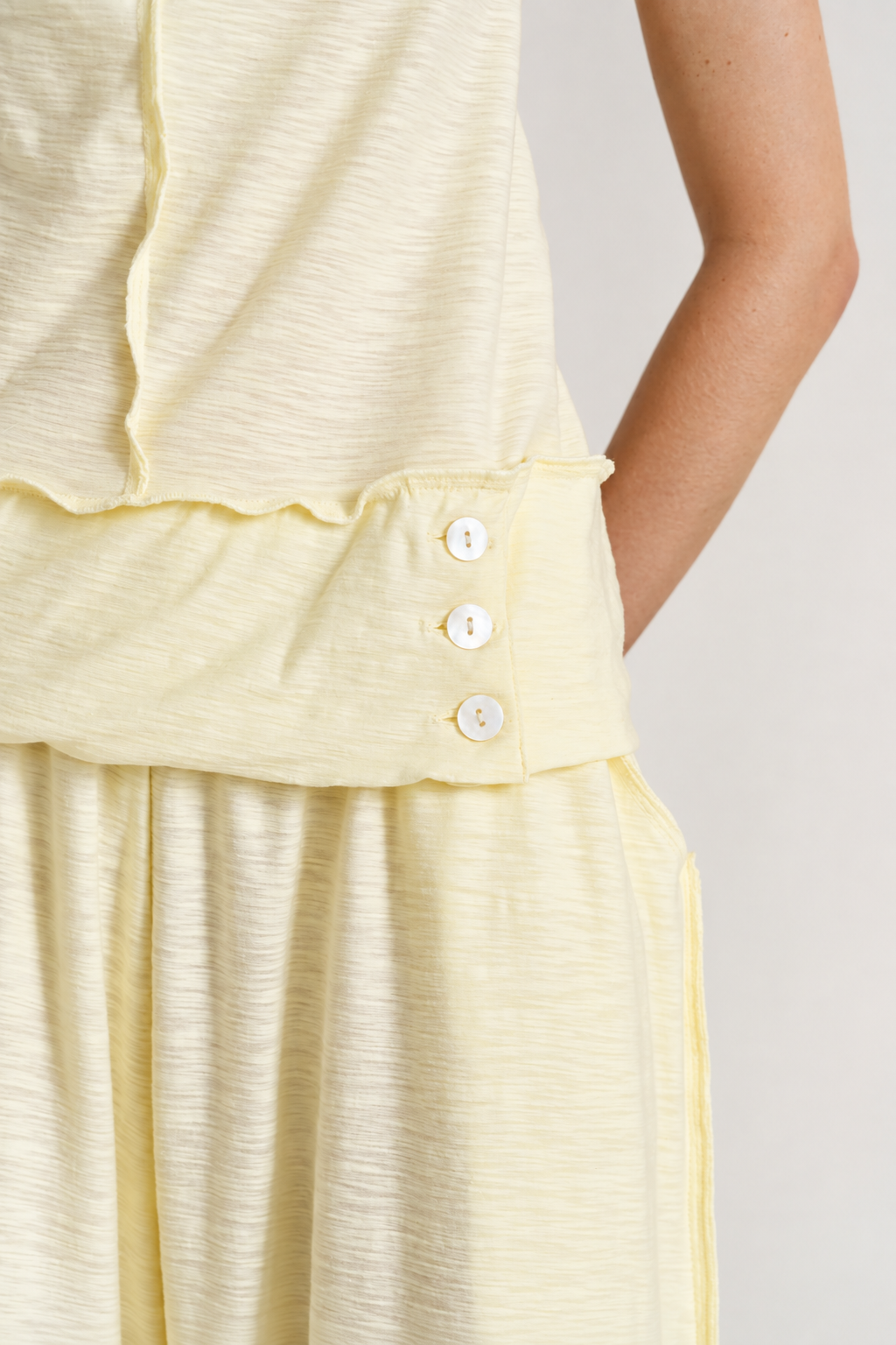Close-up of a pale yellow sleeveless top or dress with a ruffled waistband and three white buttons, shown on a model against a light neutral background.