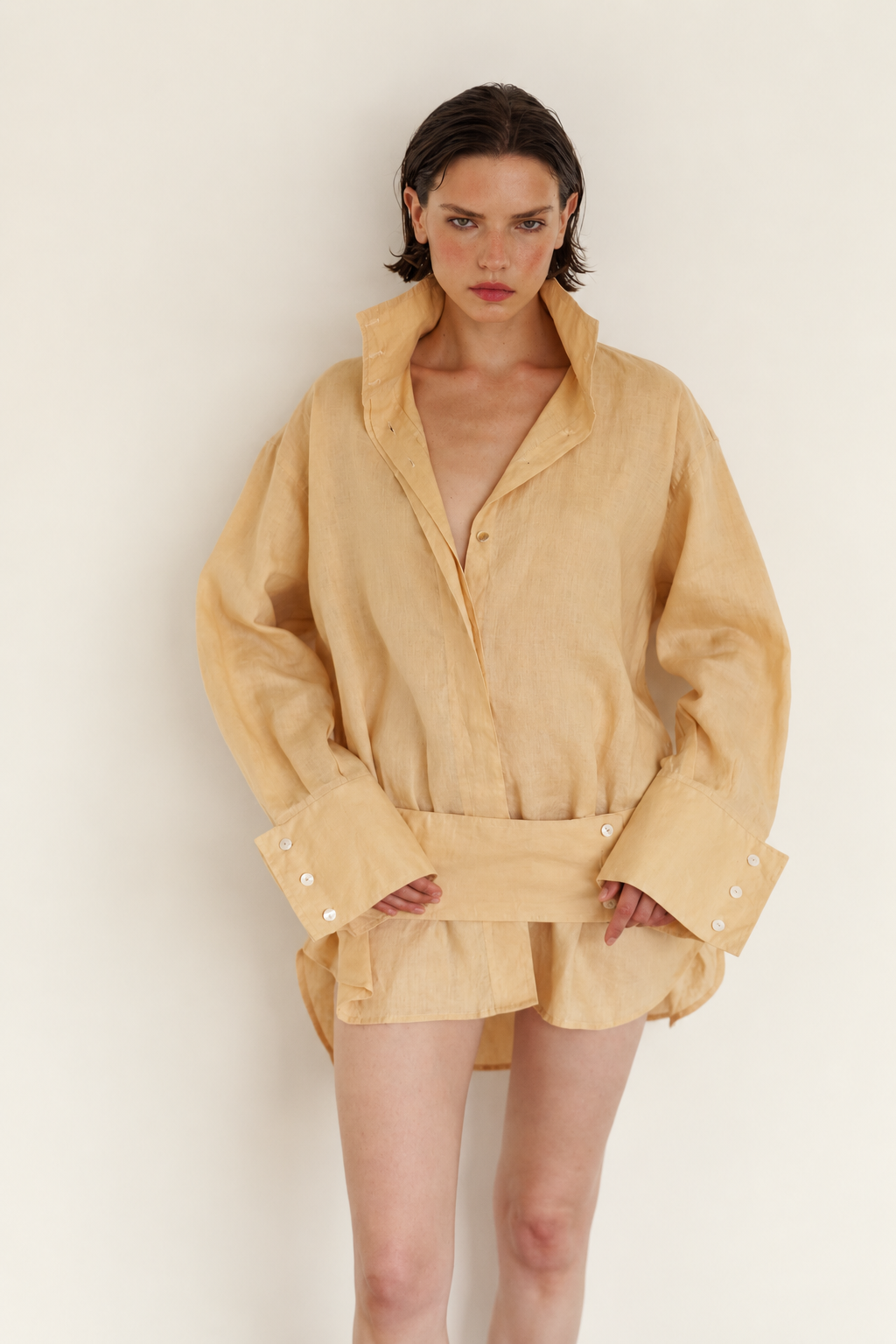 A model wearing a light beige linen shirt jacket with a shawl collar, belted waist, and buttoned cuffs, set against a plain off-white background.