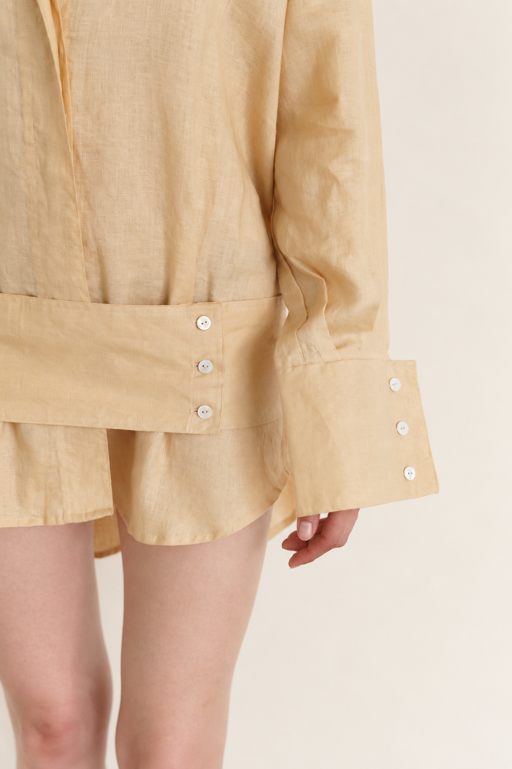 Close-up of a person wearing a light beige linen shirt dress with a belted waist and buttoned cuffs, showing the fabric texture and details.