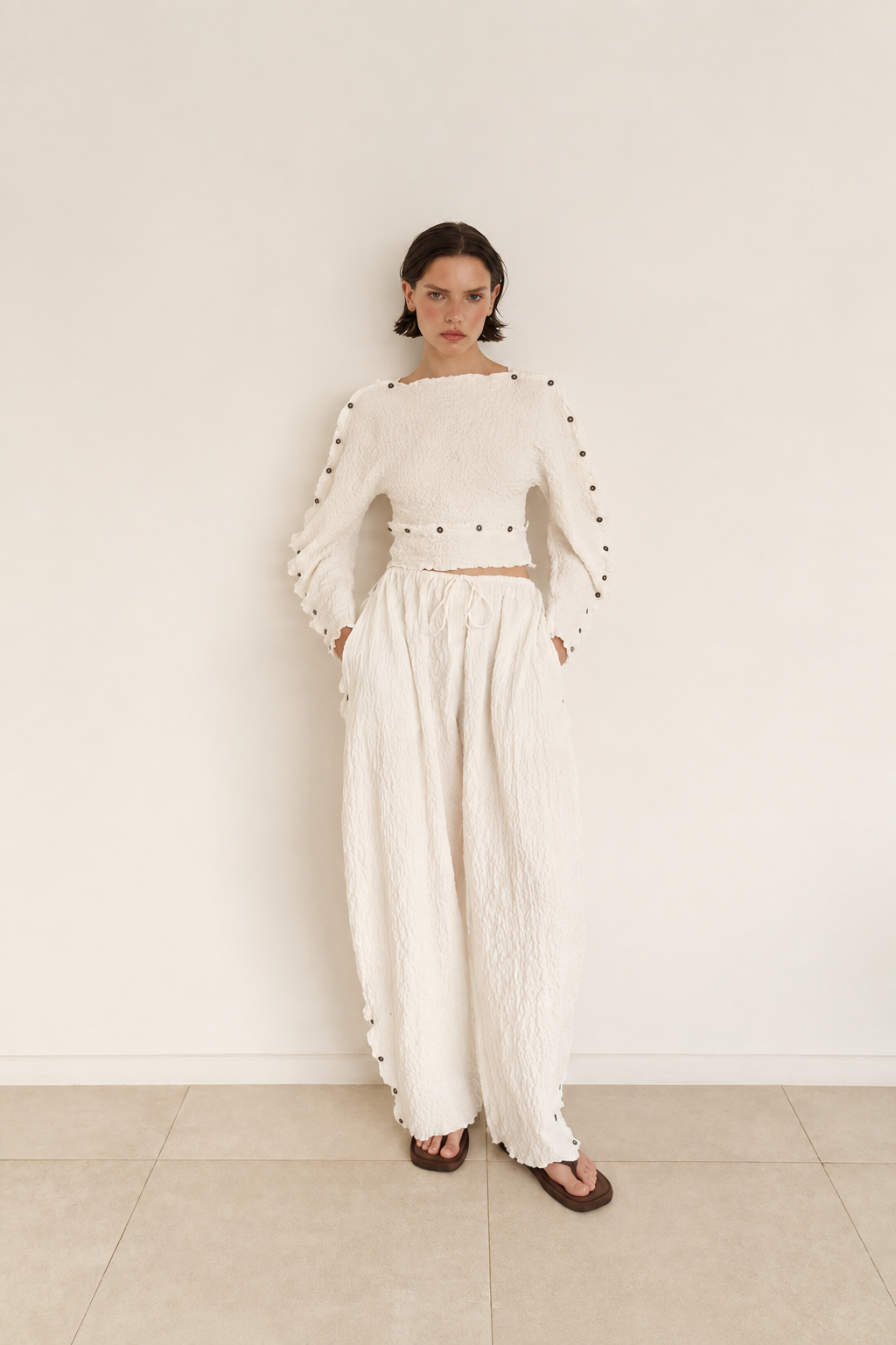 A model stands against a plain off-white wall wearing a matching cream-colored two-piece outfit consisting of a cropped long-sleeve top with studded details on the sleeves and wide-leg textured pants, paired with dark brown sandals.