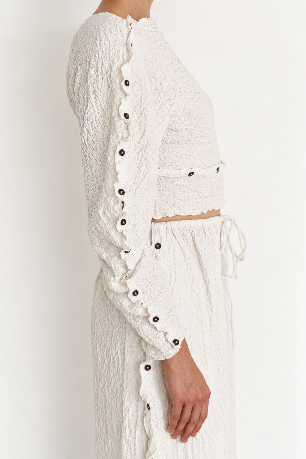 Woman wearing a white crinkle set with shell buttons on the side of the sleeve and side of the pants. The waist also has buttons and the pants is closed with a drawstring. 