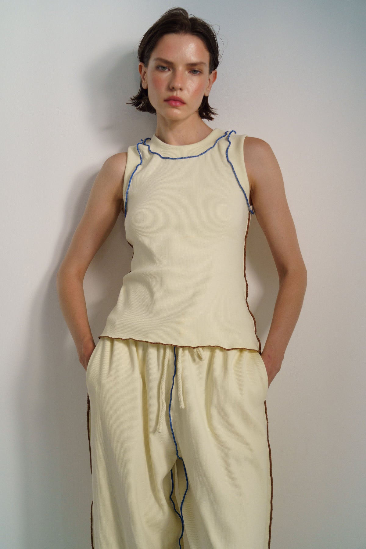 A model wearing a cream-colored sleeveless knit top and matching drawstring pants with thin blue and brown piping details, standing against a plain light gray wall.