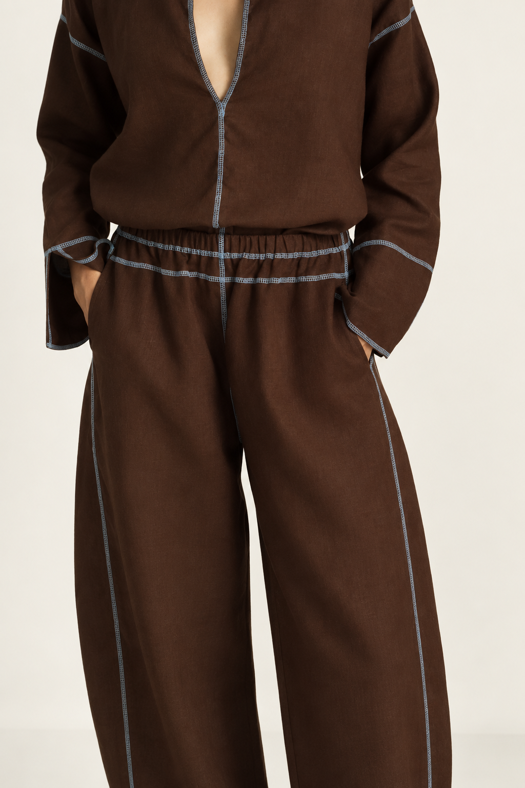 A close-up of a model wearing a matching brown two-piece set with light blue contrast piping, featuring a long-sleeve top with a deep V-neck and wide-leg trousers.