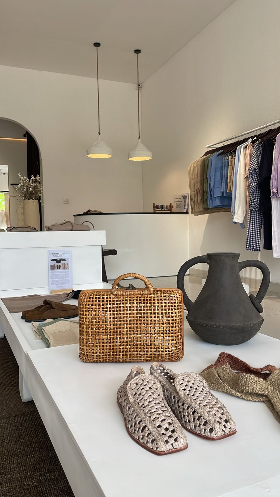 A styled interior scene showing a woven straw handbag on a white table, with crochet slip-on shoes, a dark ceramic pitcher, and clothing hanging in the background.