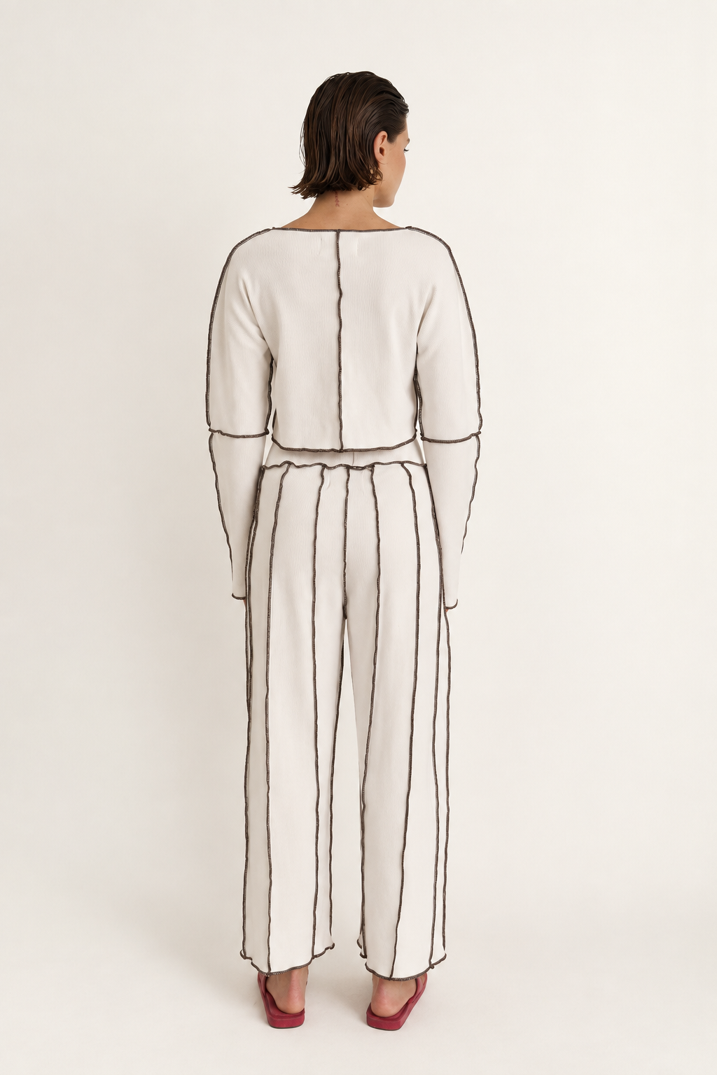 A model viewed from the back wearing a cream-colored two-piece set with black embroidered vertical lines and a matching waist tie, paired with red heels, against a plain off-white background.